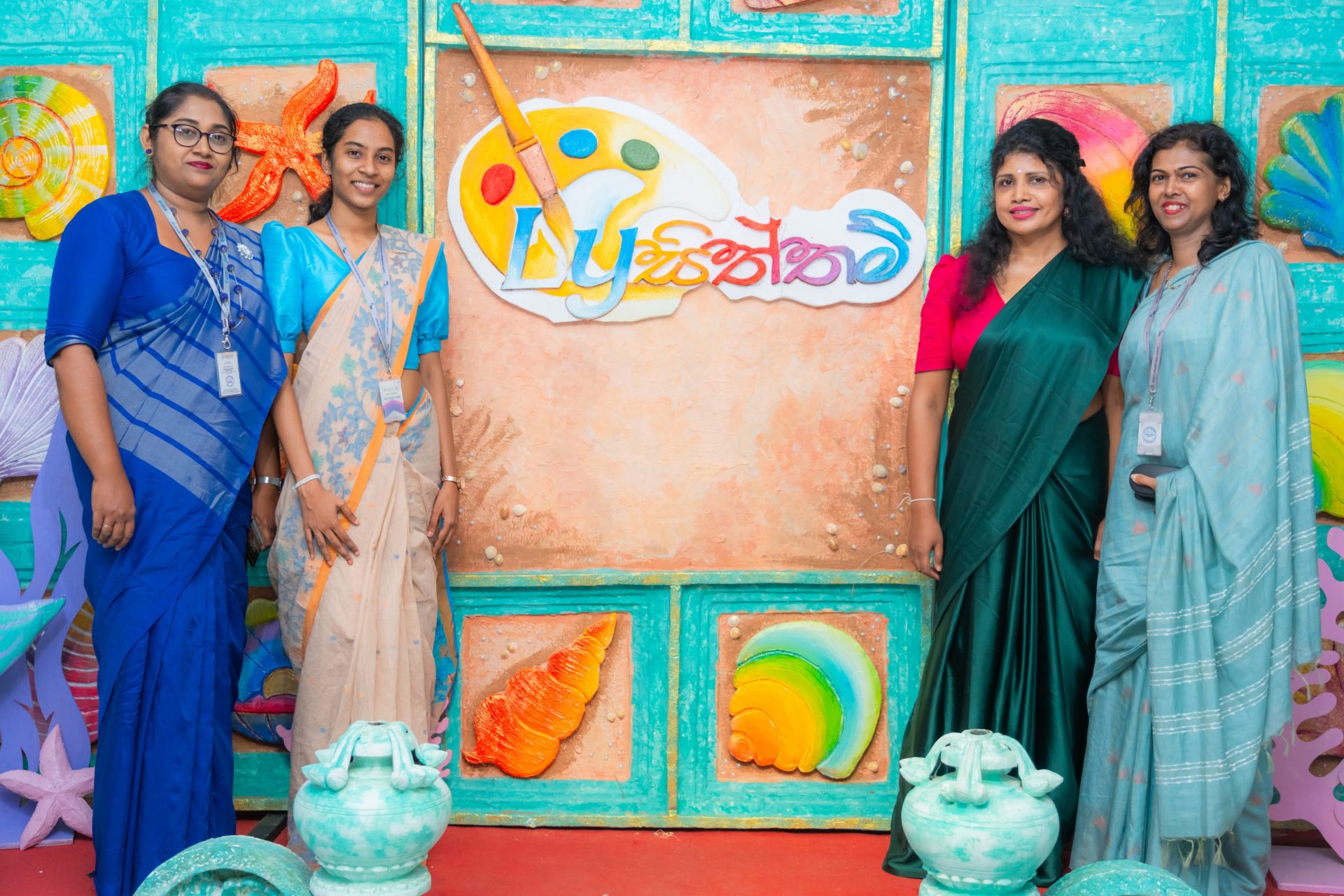 Four women in sarees pose at a colorful, marine-themed art festival with creative decorations.