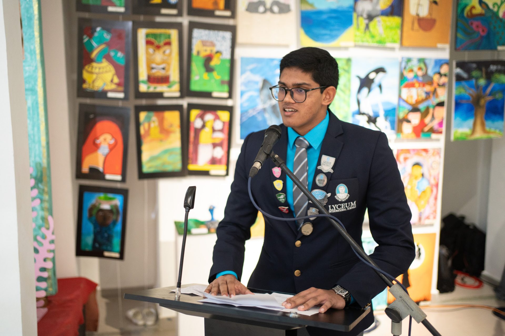 Student leader speaks at Lyceum International School art event with colorful student artwork in background.