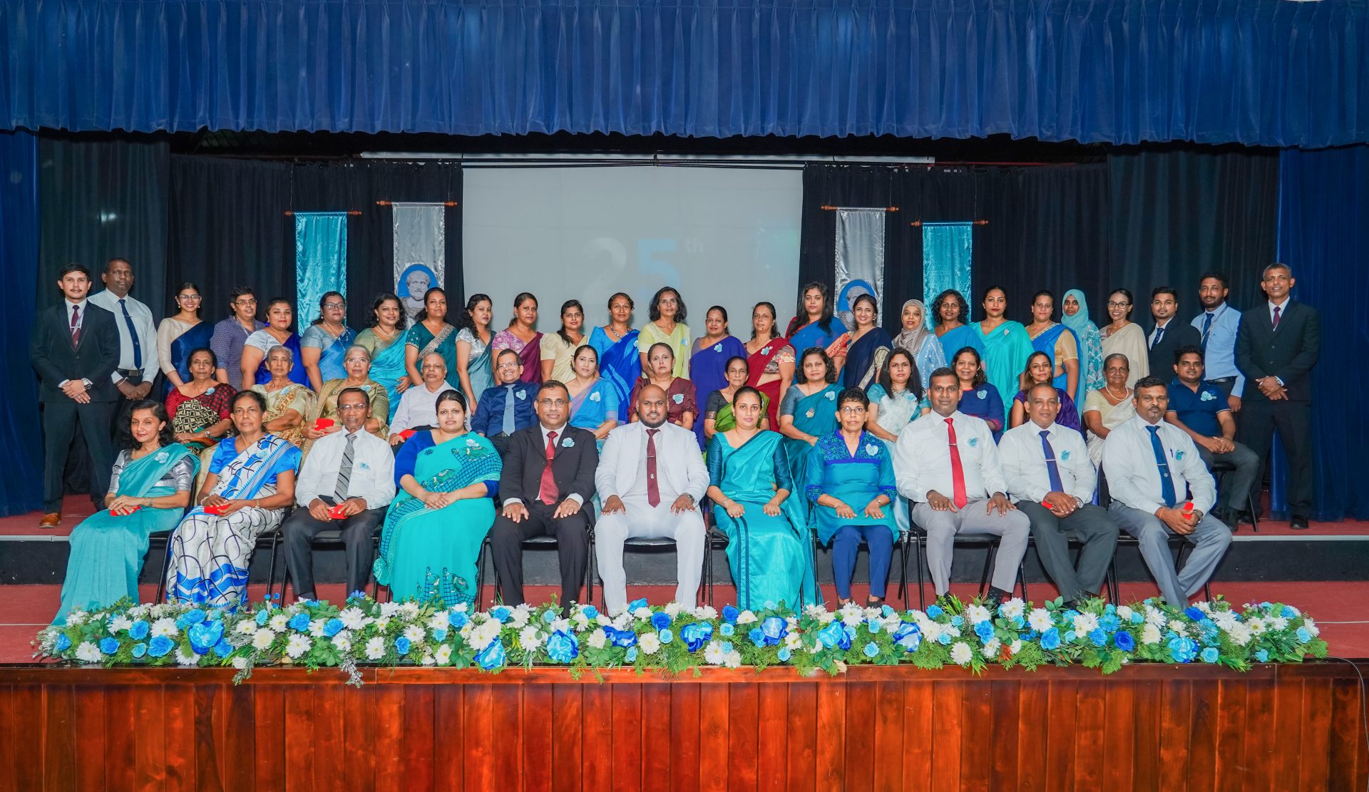 Staff Group Photo on Stage for 32nd Anniversary Celebration Staff group photo on stage celebrating 32nd anniversary with blue sarees, suits, banners, and flowers.