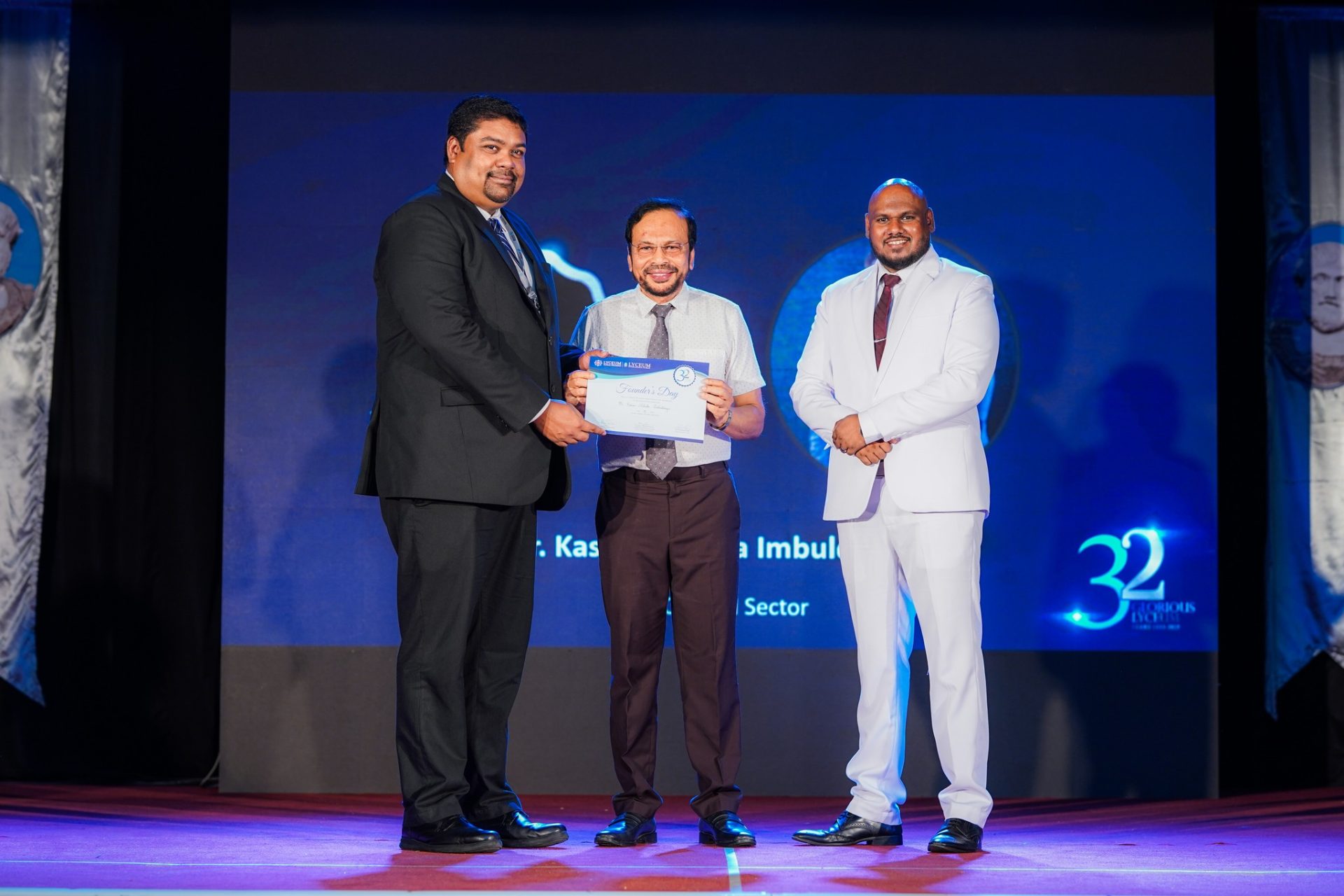 Celebrating Excellence: Award Ceremony Marks 32nd Anniversary Award ceremony on stage with three men, certificate presentation, blue anniversary theme, and official banners.