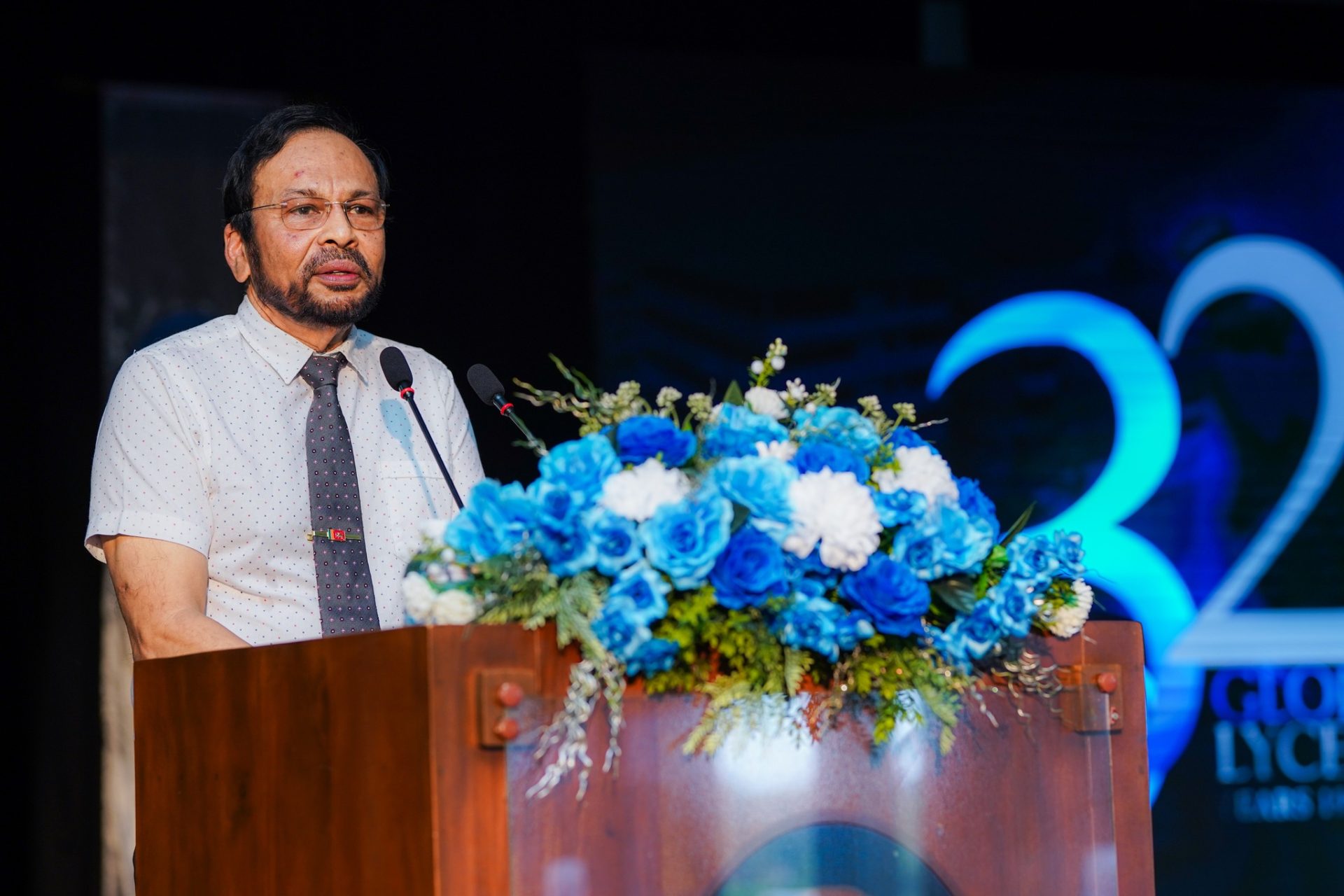 Speaker Addresses 32nd Anniversary Event with Floral Podium Man giving speech at podium with blue flower arrangement during 32nd anniversary celebration event.