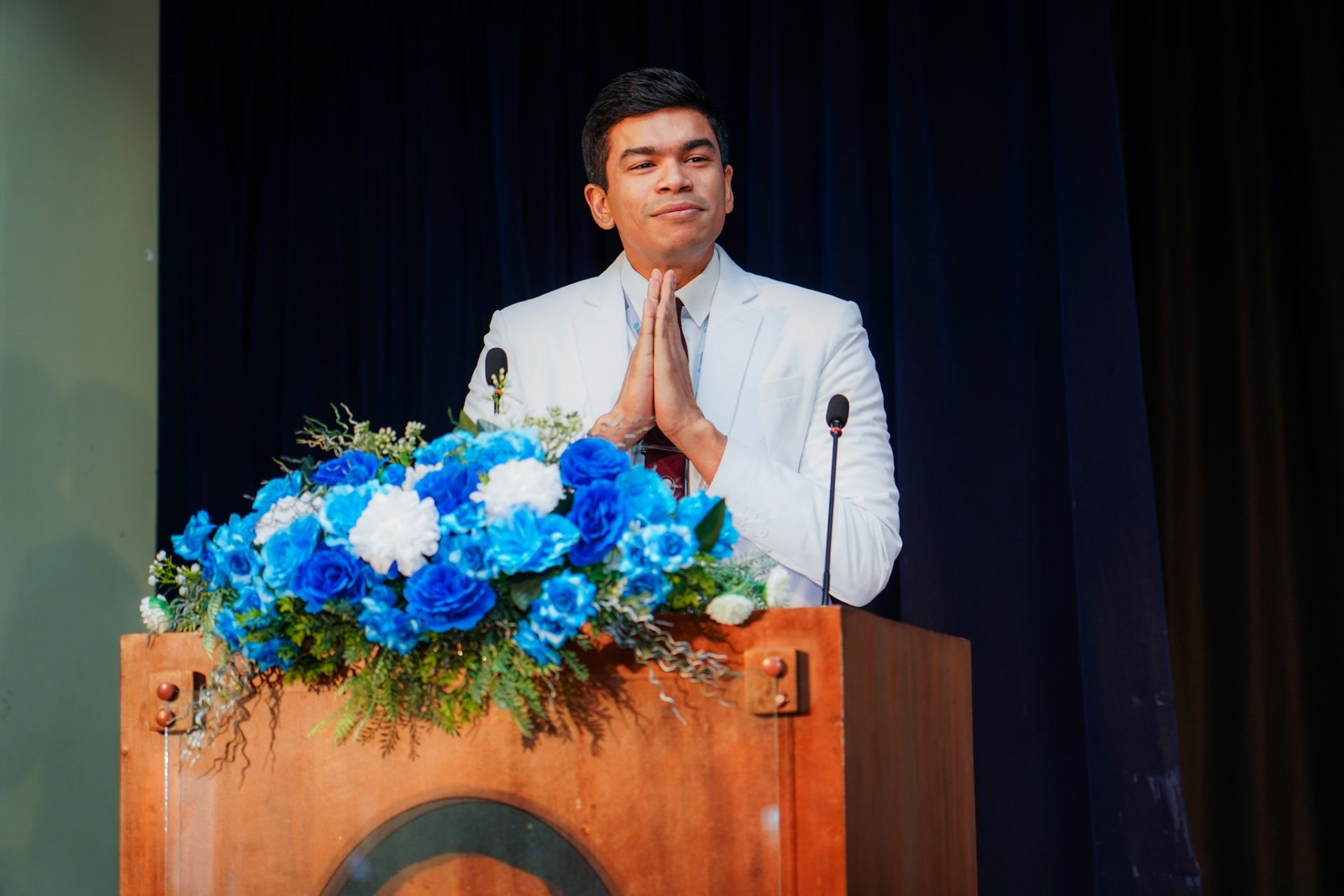 Man in White Suit Gives Speech at Floral-Adorned Podium Man in white suit speaking at podium with blue floral arrangement at formal event.