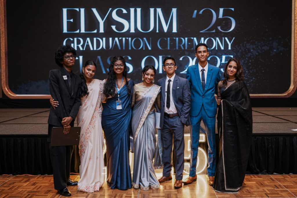 Group of graduates in sarees and suits posing at Elysium 2025 graduation ceremony stage.
