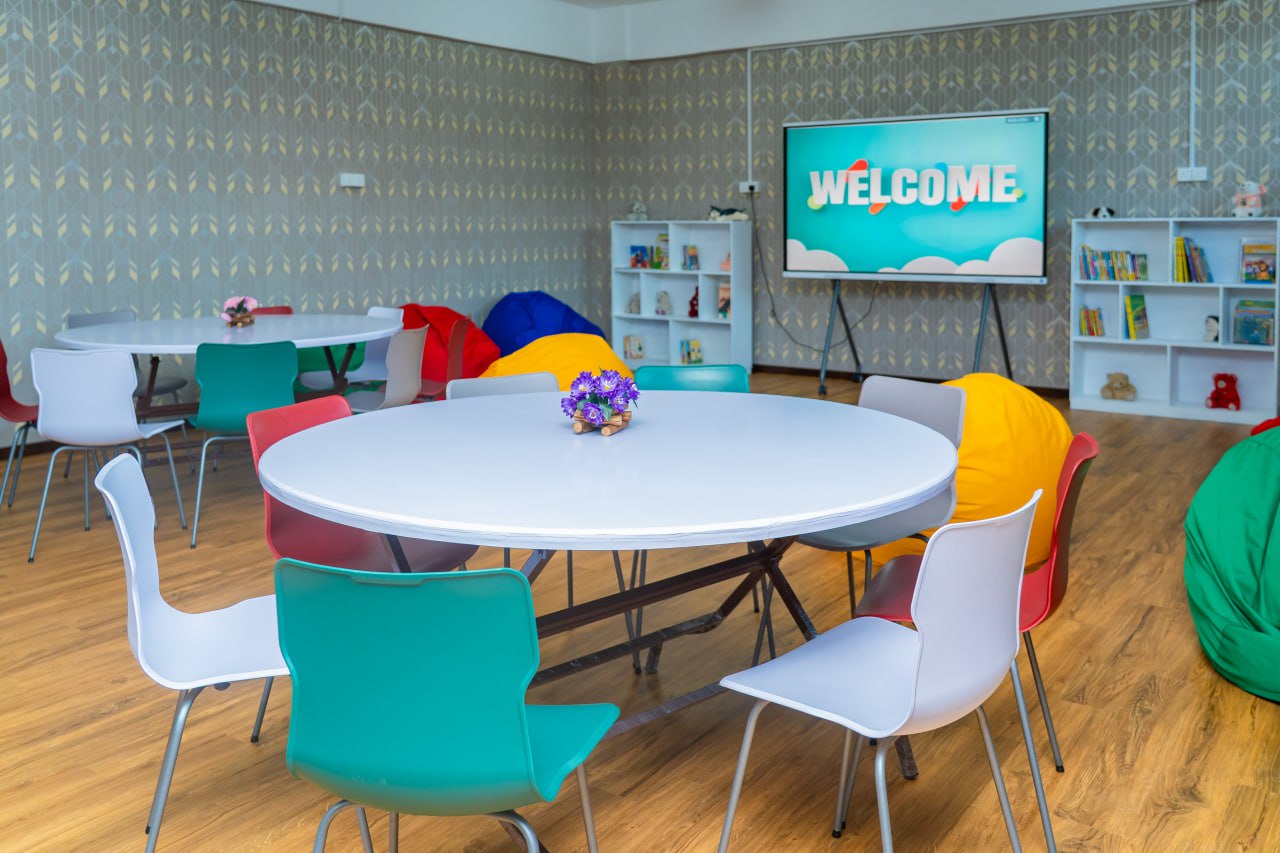Colorful kids classroom with round tables, bean bags, bookshelves, and a welcome screen for activities.