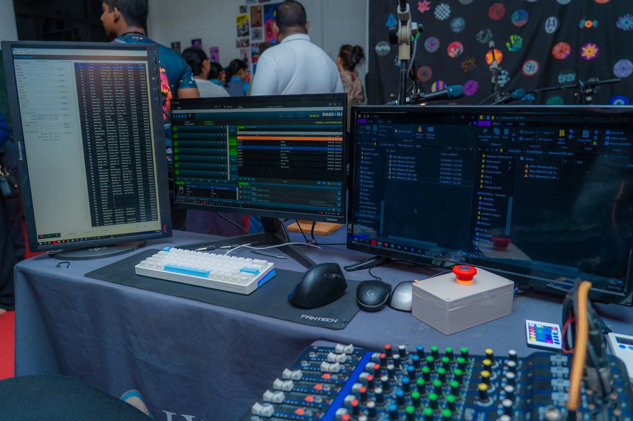 Broadcast studio workstation with audio mixers, multiple monitors, emergency stop button, and event attendees.