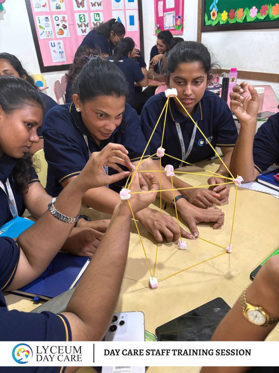 Daycare staff team-building activity with spaghetti and marshmallows during training session in colorful classroom.