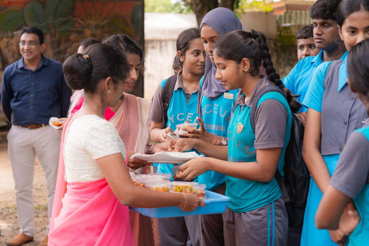 Students in school uniforms receiving food from teachers during a cultural celebration outdoors.