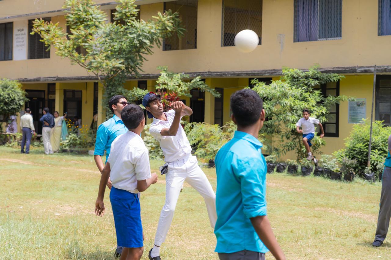 School boys playing volleyball on campus lawn during break, supervised by teachers, sunny day.