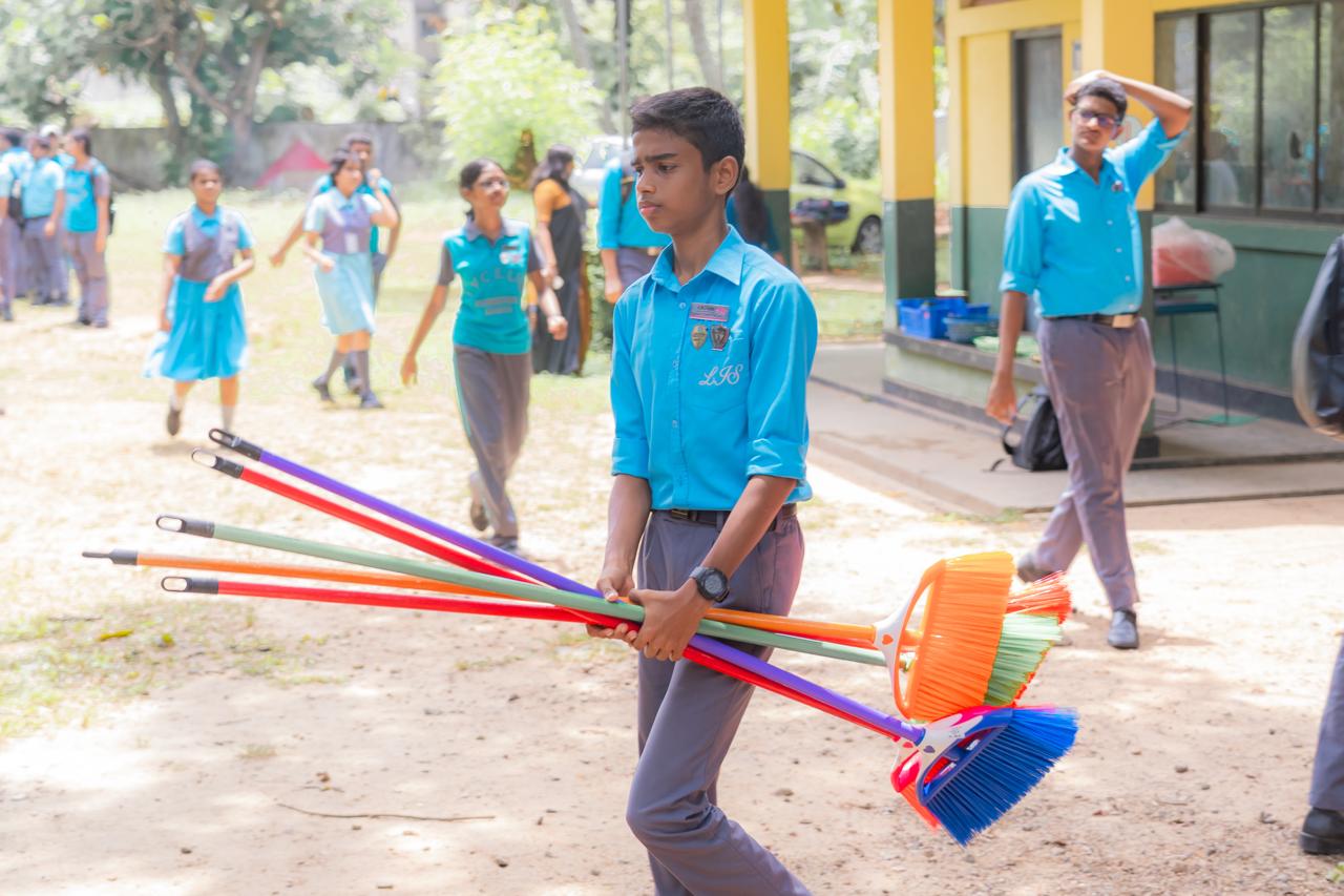 School children in uniforms carrying colorful brooms during a campus cleaning activity outdoors.