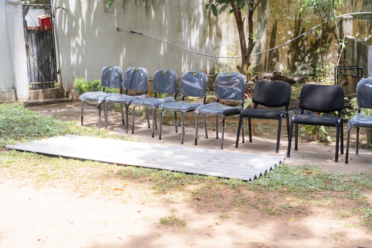 Row of plastic-wrapped chairs and construction materials outdoors near concrete wall and tree in sunlight.
