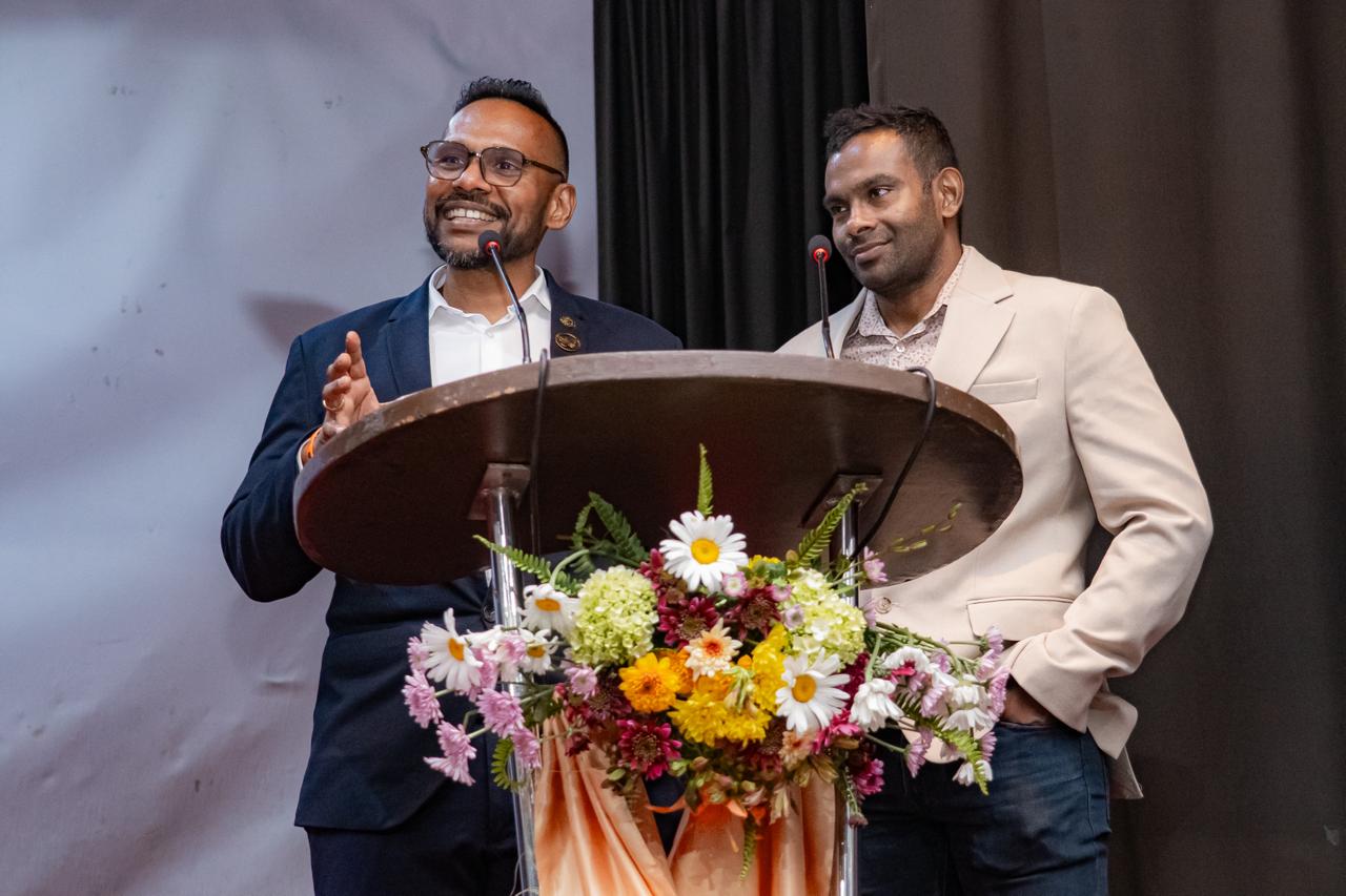 Two men speak at a flower-adorned podium during a formal conference or award event.