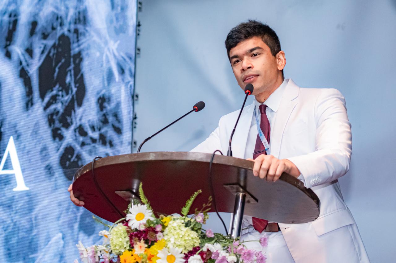 Man in white suit delivers keynote speech at podium with floral arrangement and abstract digital background.
