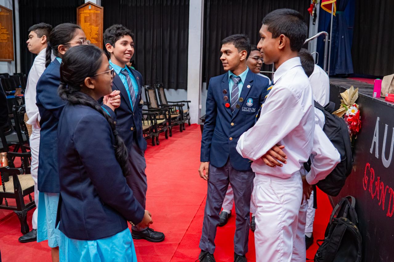 High school students in formal uniforms chatting at academic ceremony in auditorium with red carpet.