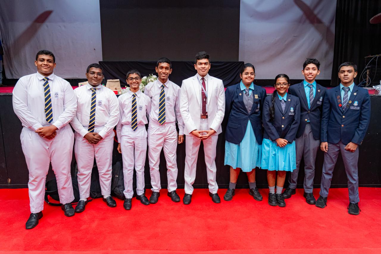 Group of students in formal school uniforms at awards ceremony on red carpet indoors.