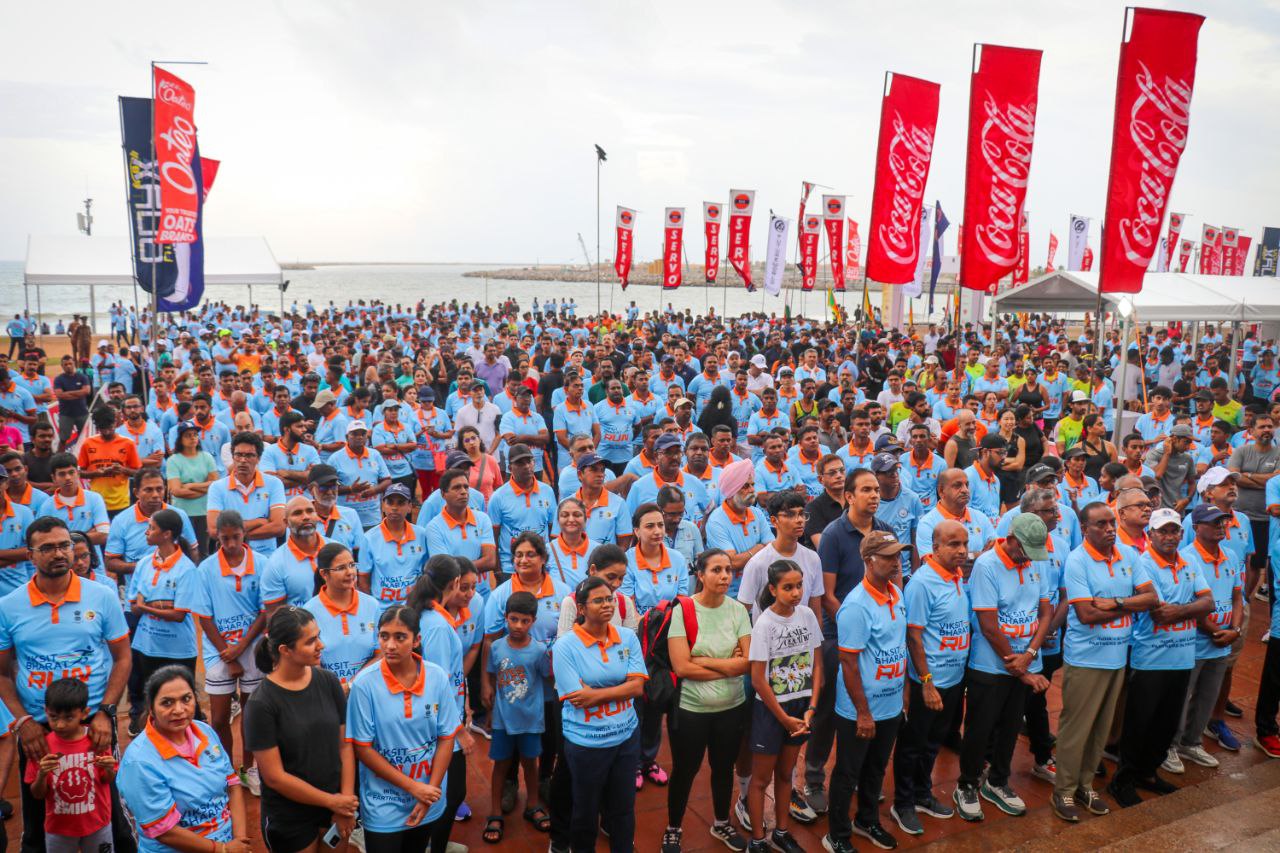 Large crowd at Viksit Bharat Run event near waterfront with branded banners and blue t-shirts.