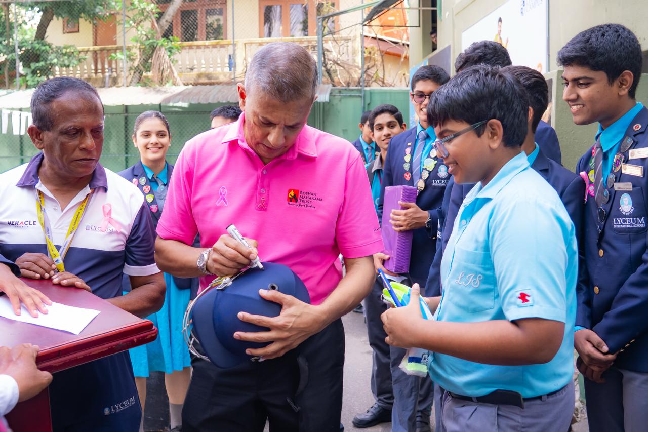 Guest signing helmet at Lyceum International School breast cancer awareness event with students and staff.