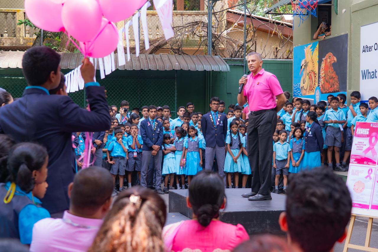 School breast cancer awareness event with students in pink, speaker, balloons, and educational posters outside.