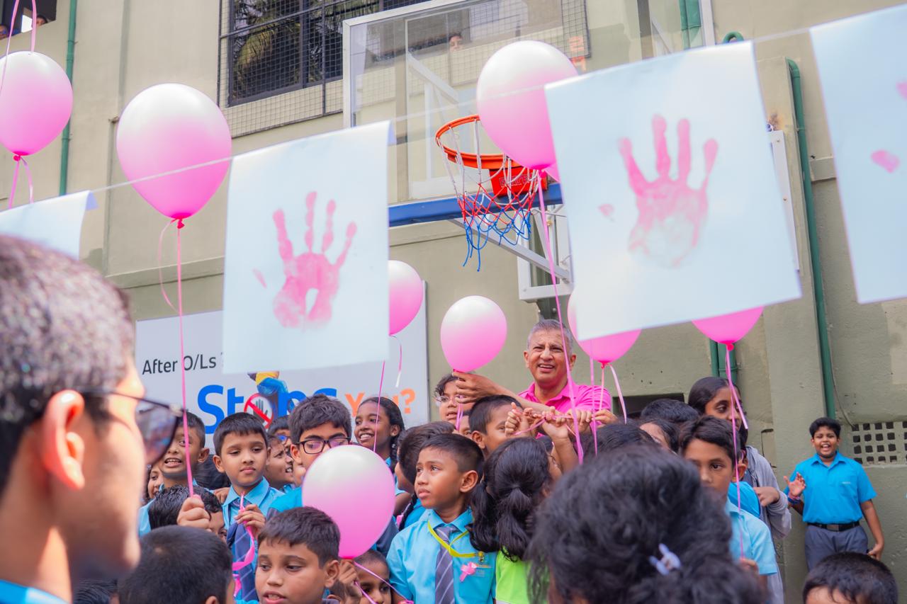 School children holding pink balloons and handprint signs at an anti-bullying awareness event outdoors.