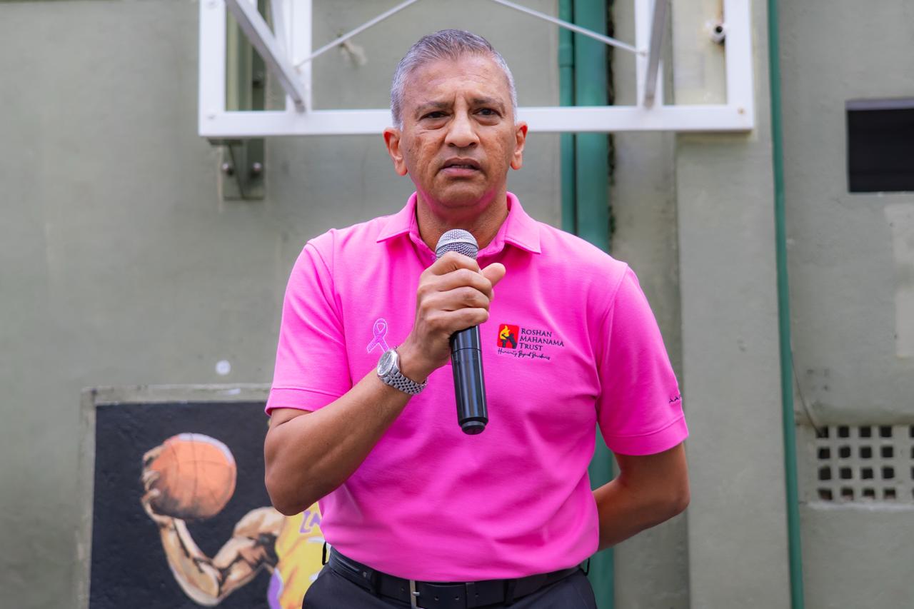 Man in pink Roshan Mahanama Trust shirt speaks at sports charity event for breast cancer awareness.