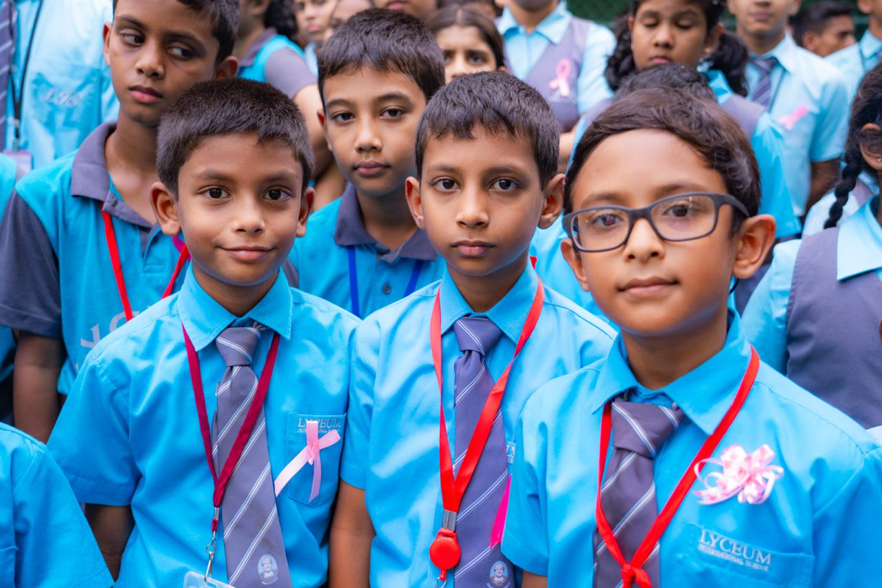 School children in blue uniforms with pink ribbons at outdoor assembly for awareness event.
