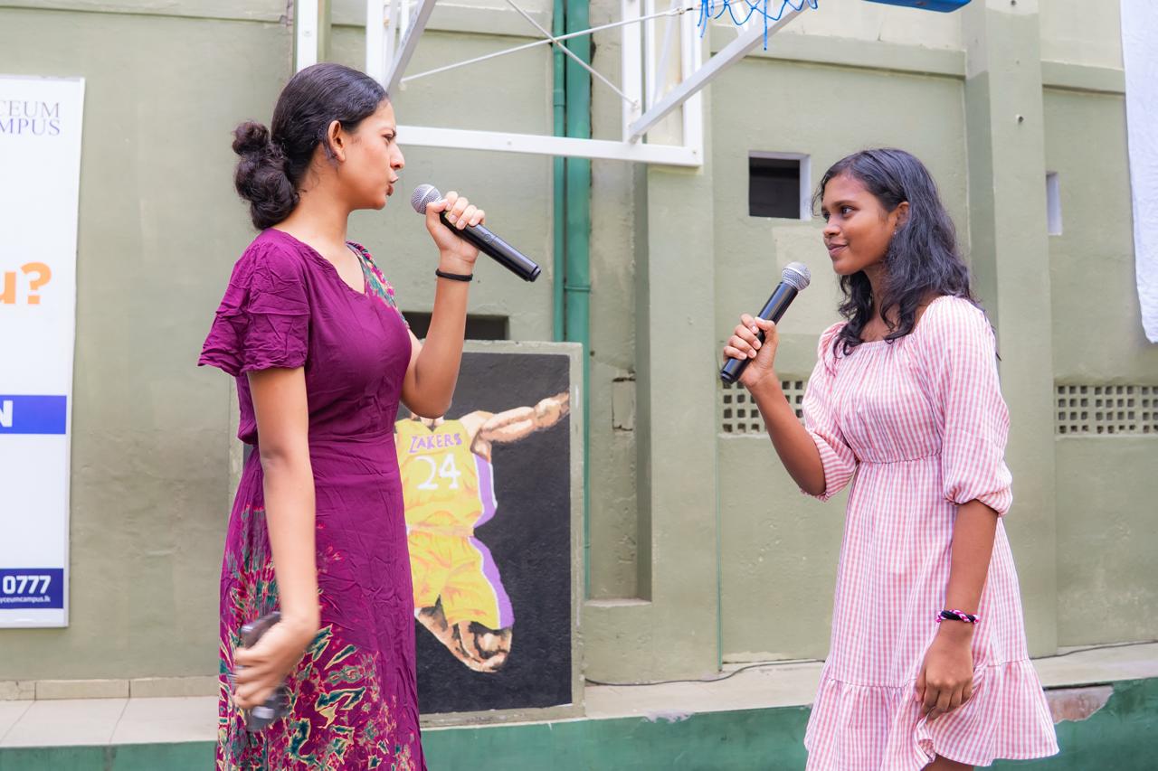 Two young women with microphones debate or perform duet at school event near Kobe Bryant mural.