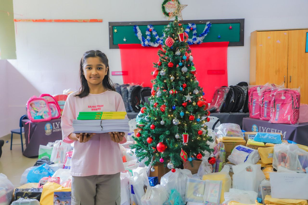 Student donates school supplies by Christmas tree, surrounded by gift bags, for charity at lyceum.
