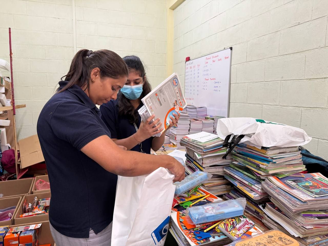 Women packing school supplies for donation, with books and stationery on table, community service project.