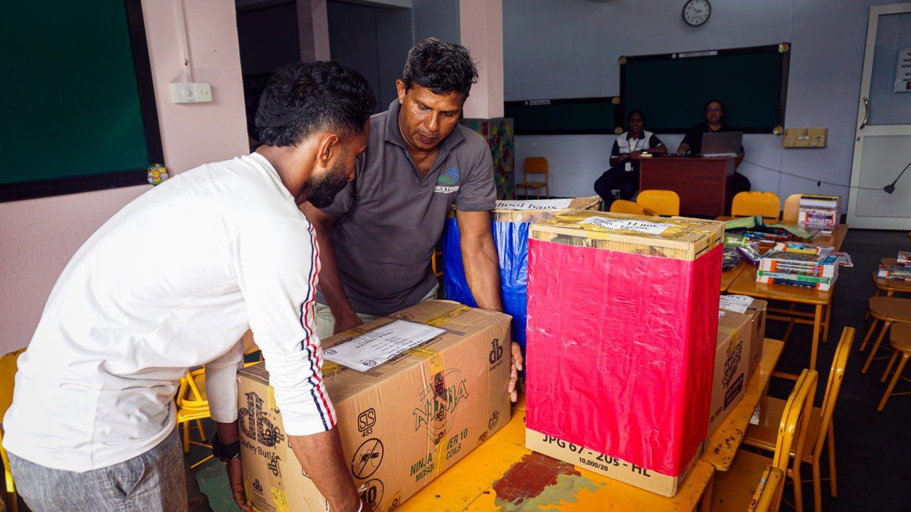 Teachers unpacking classroom supply boxes in a school, yellow chairs and chalkboard visible.