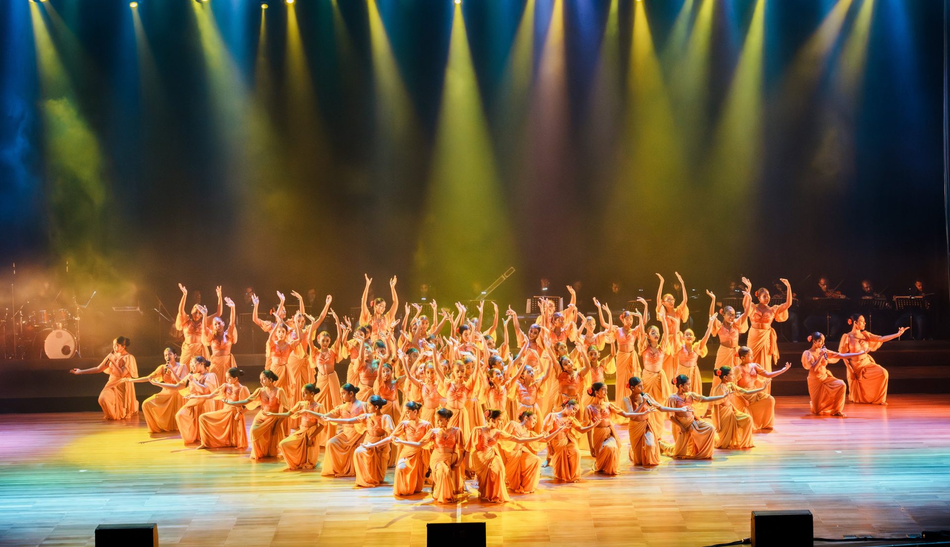 Large group of dancers in orange costumes performing synchronized choreography on a brightly lit stage.