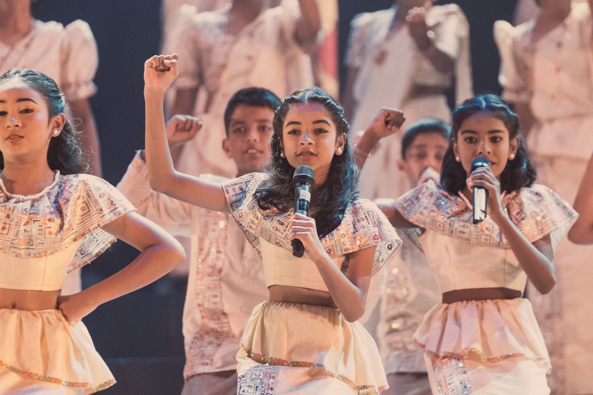 Children in traditional costumes perform energetic group song and dance on stage at cultural event.