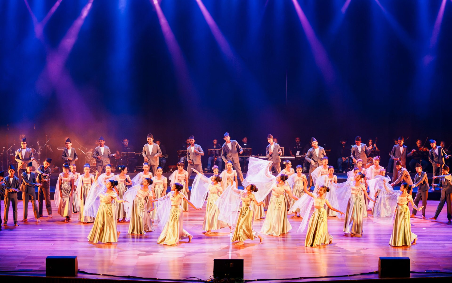 Stage performance with dancers in gold gowns, male performers in formal wear, live band, dramatic lighting.
