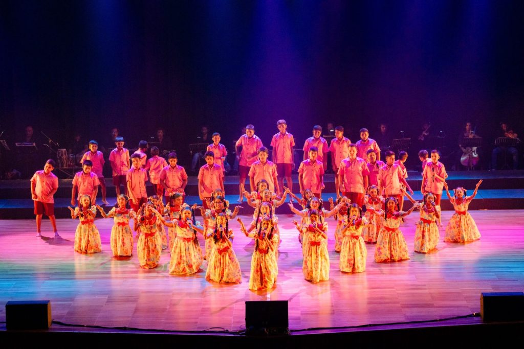 Children perform traditional folk dance on stage in colorful costumes with live music in the background.
