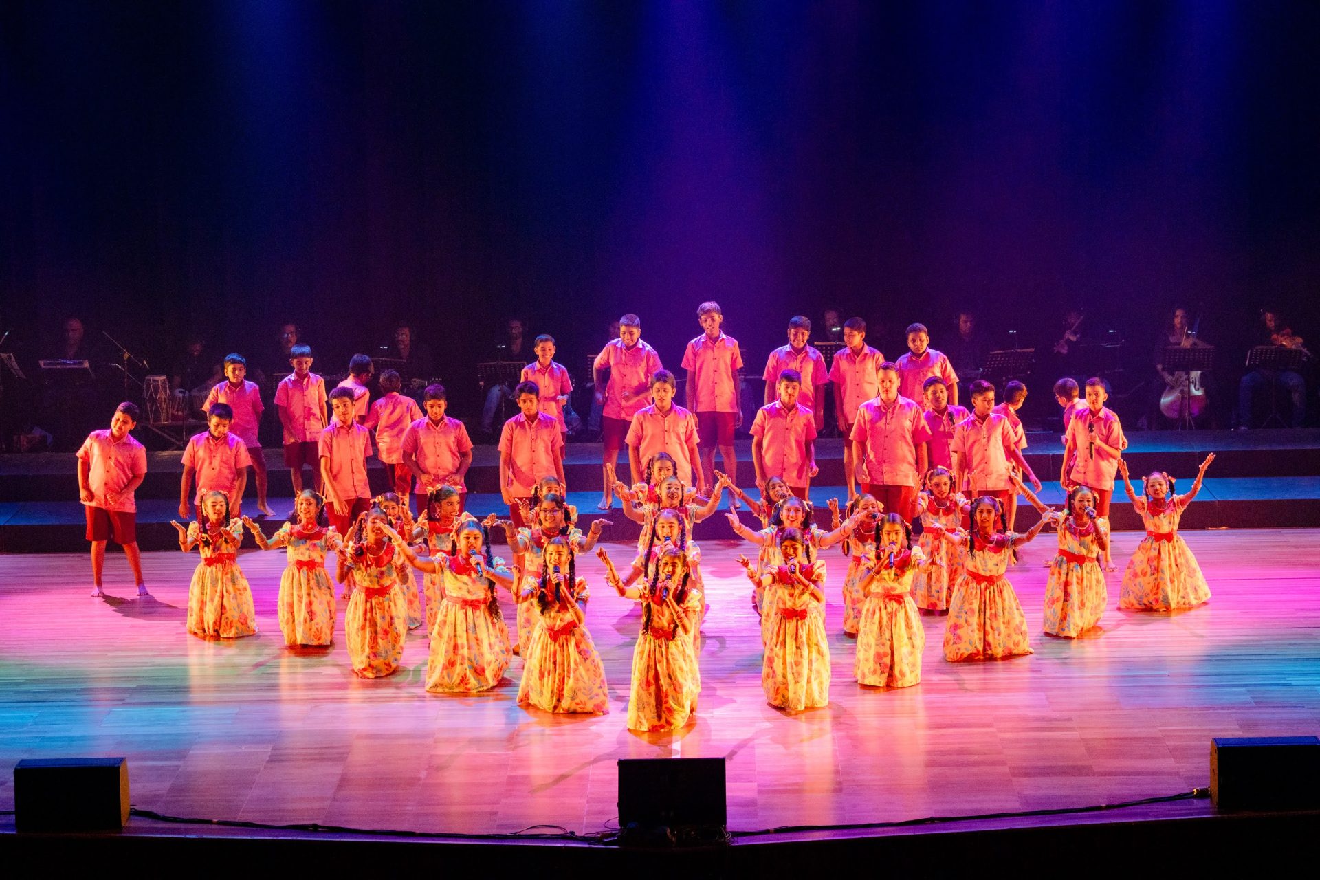 Children perform traditional folk dance on stage in colorful costumes with live music in the background.