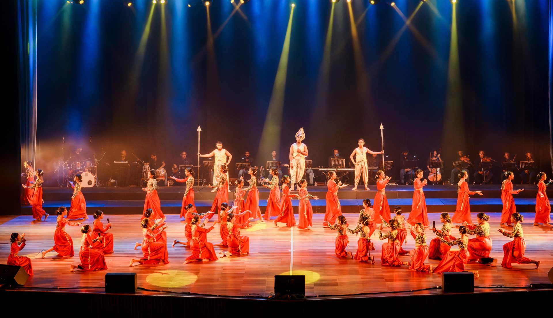 Colorful traditional stage dance performance with live musicians, red costumes, and dramatic lighting.