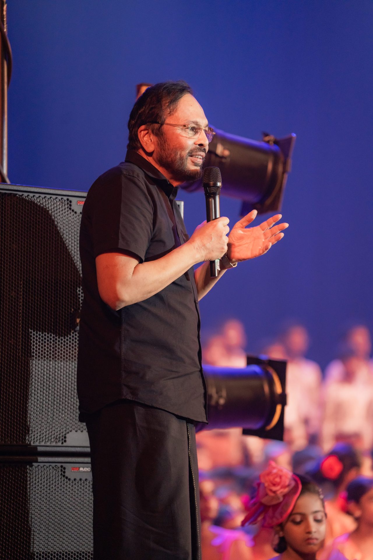 Man in black kurta speaking on stage at cultural event with children performers in background.