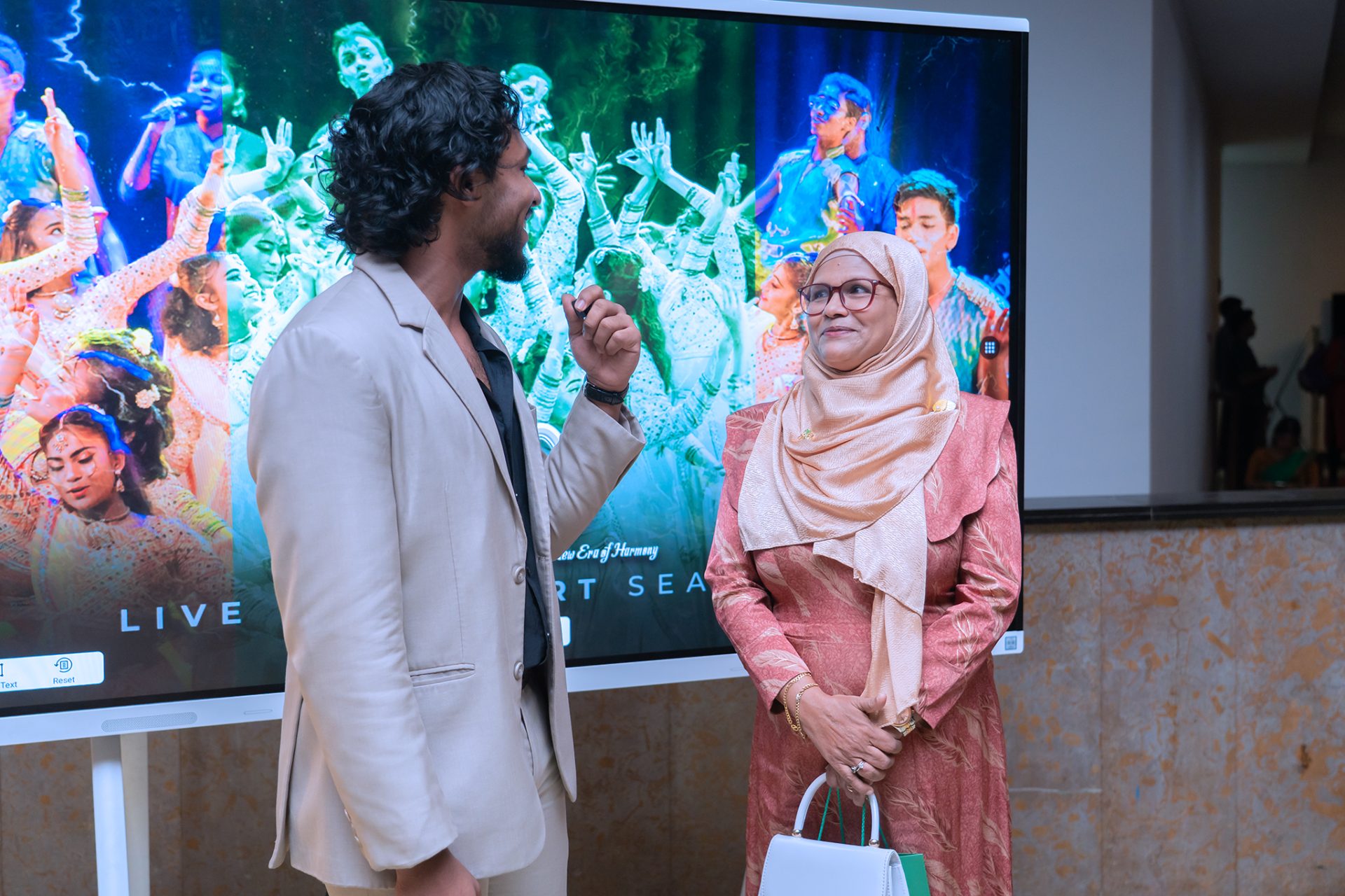 Man interviews woman in hijab at colorful cultural event with performers on screen in background.