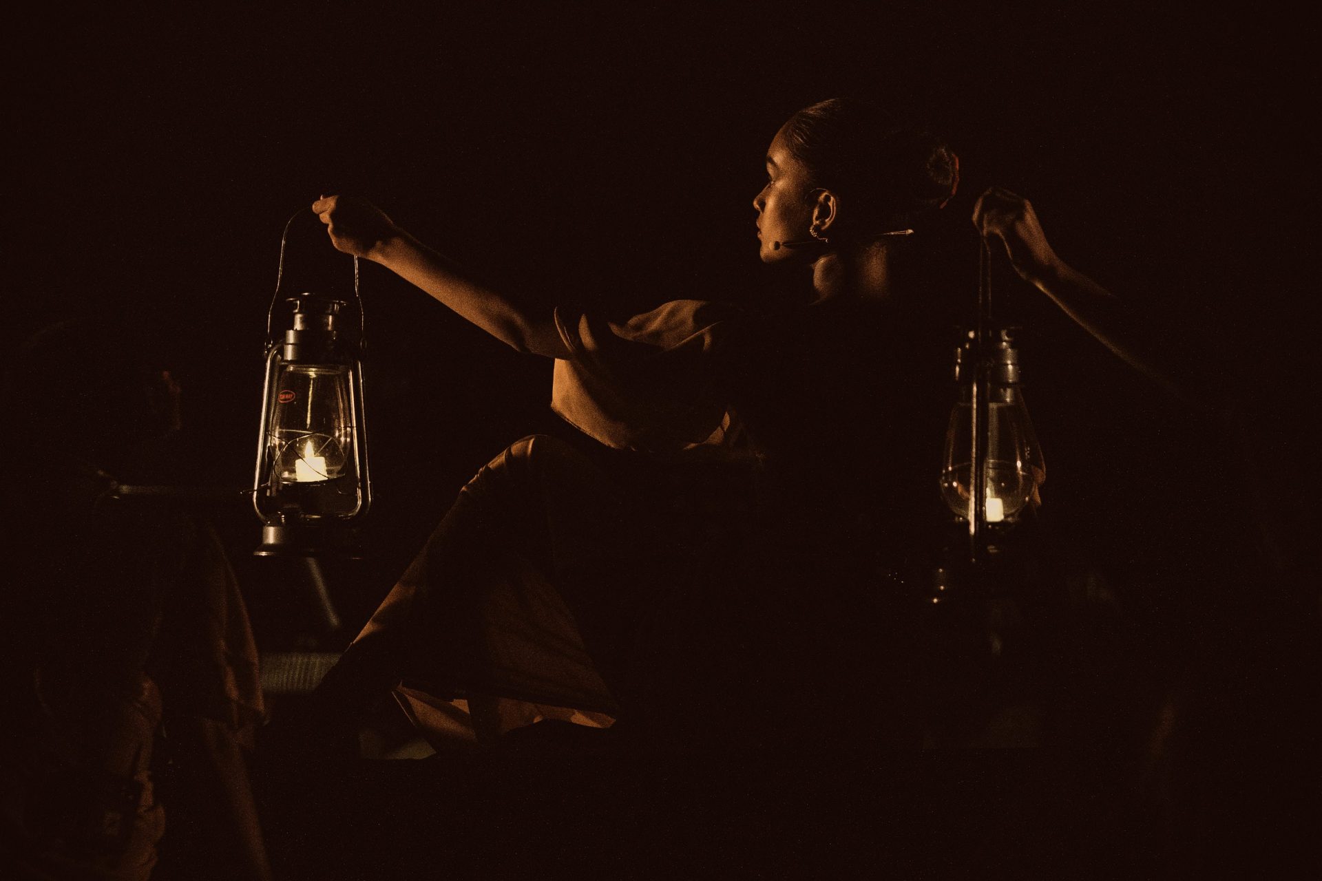 Woman holding lanterns on dark stage, dramatic lighting, mysterious performance art, night storytelling scene.