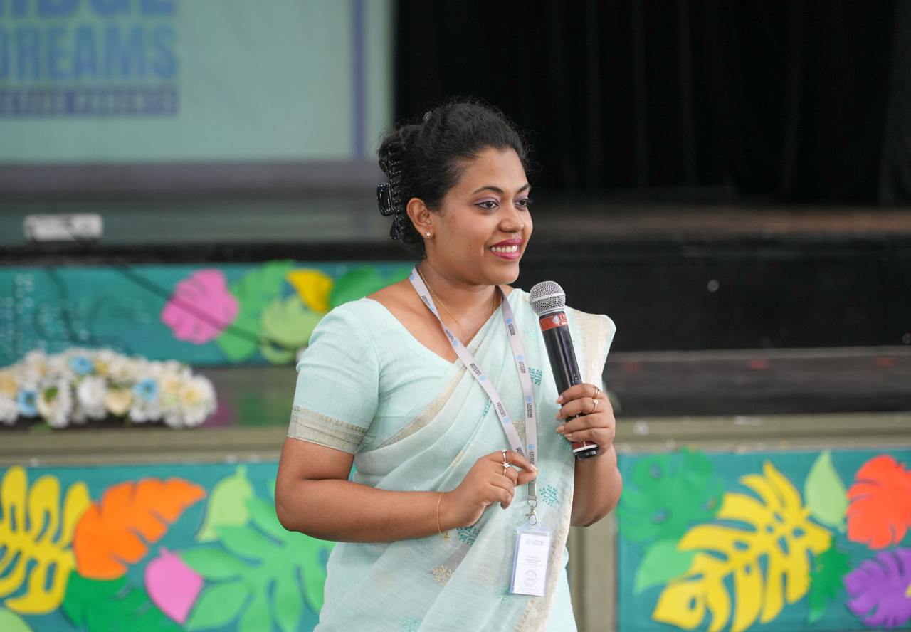 Woman in pastel saree speaks on colorful school event stage with microphone, smiling confidently.