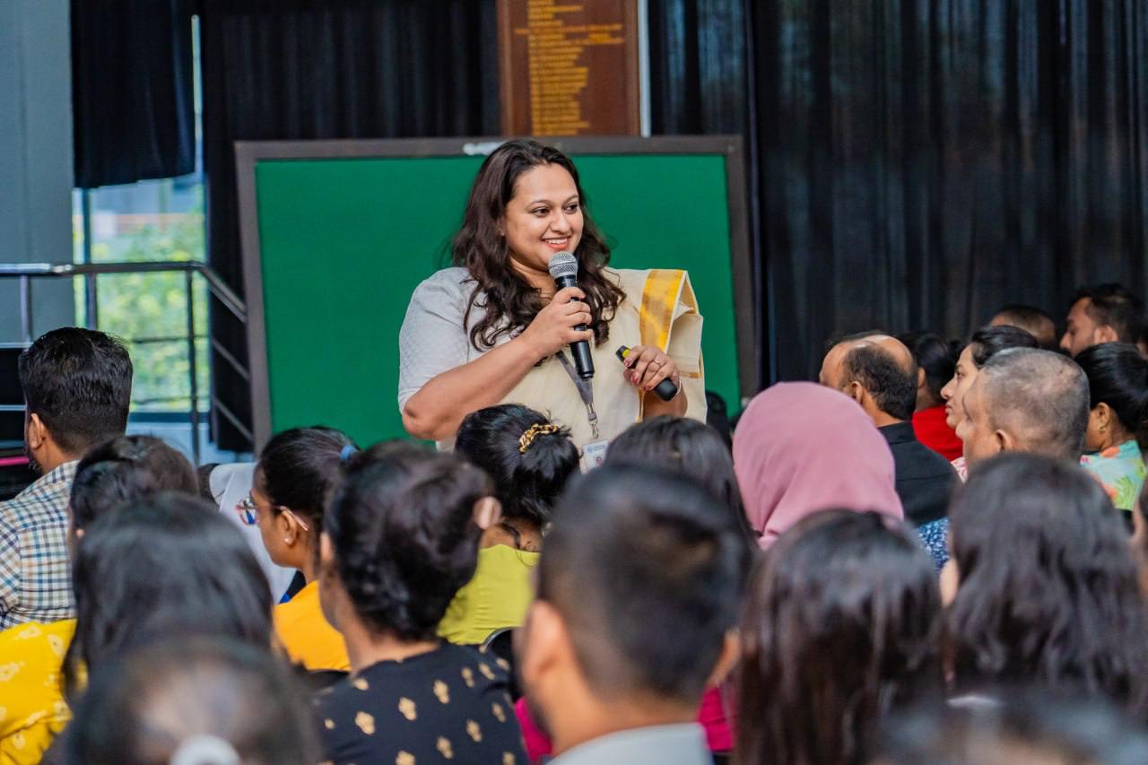 Smiling woman leads seminar, addressing diverse, attentive audience in indoor setting with green board.