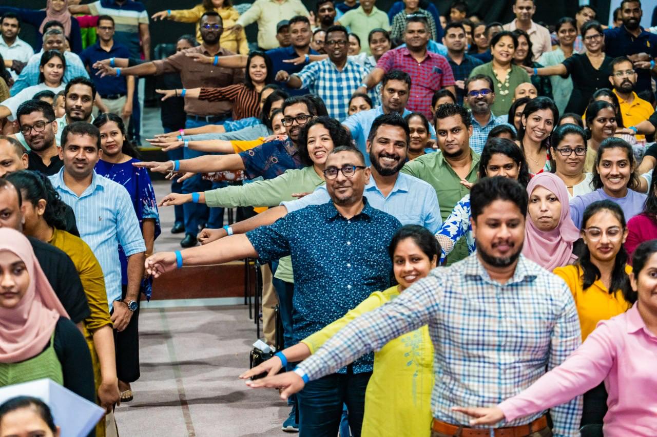 Diverse group in auditorium raising arms together during energetic team-building activity, promoting unity and inclusion.