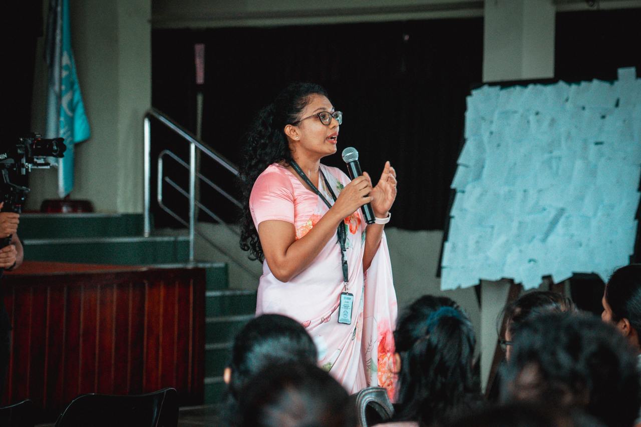 Woman in pink saree speaking at student seminar with audience listening and photographer present.
