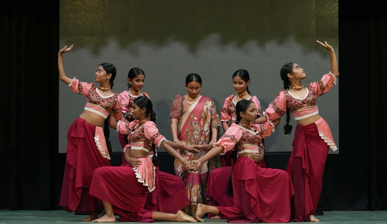 Group of girls and teacher performing synchronized Indian classical dance in vibrant traditional costumes on stage.