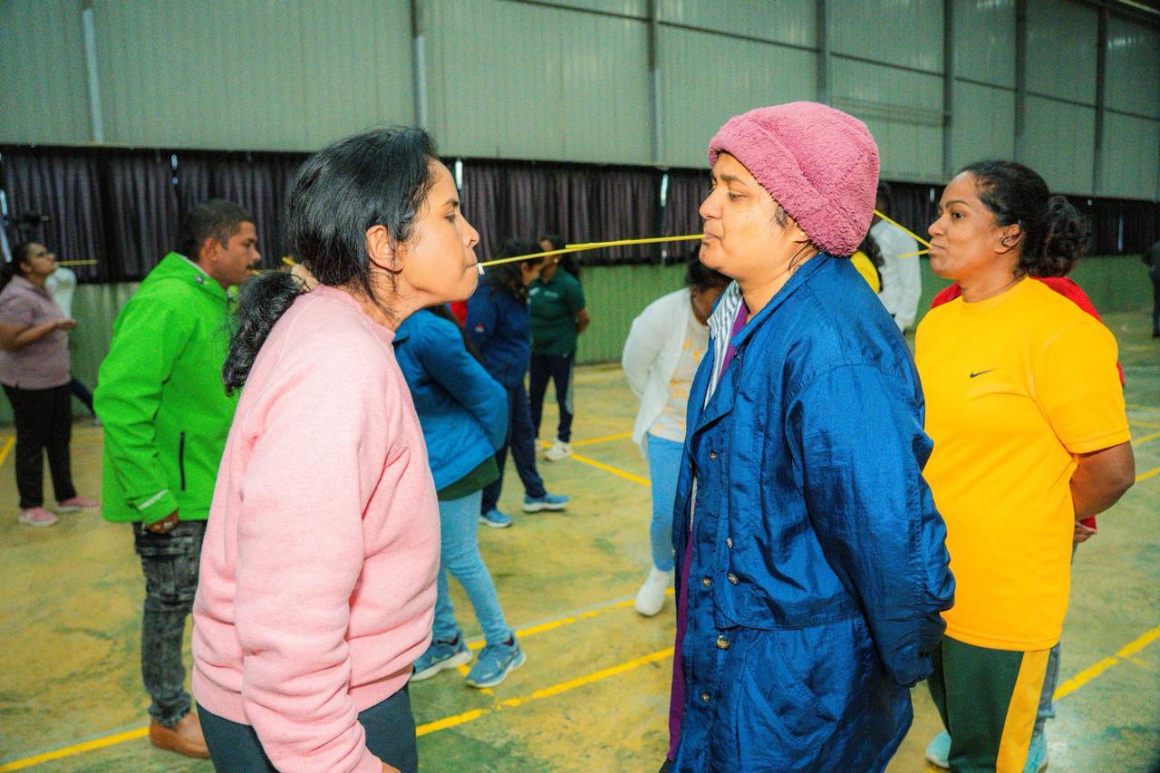 Adults balancing spaghetti sticks between mouths in a gym during a fun team-building challenge.
