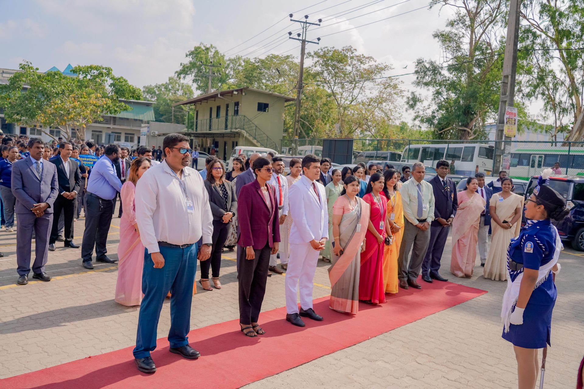 Student cadet welcomes dignitaries on red carpet at outdoor school ceremony with large audience.