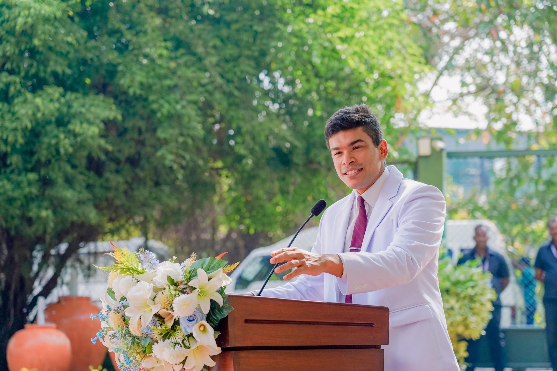 Young man in white suit speaks at outdoor ceremony podium decorated with fresh flowers.