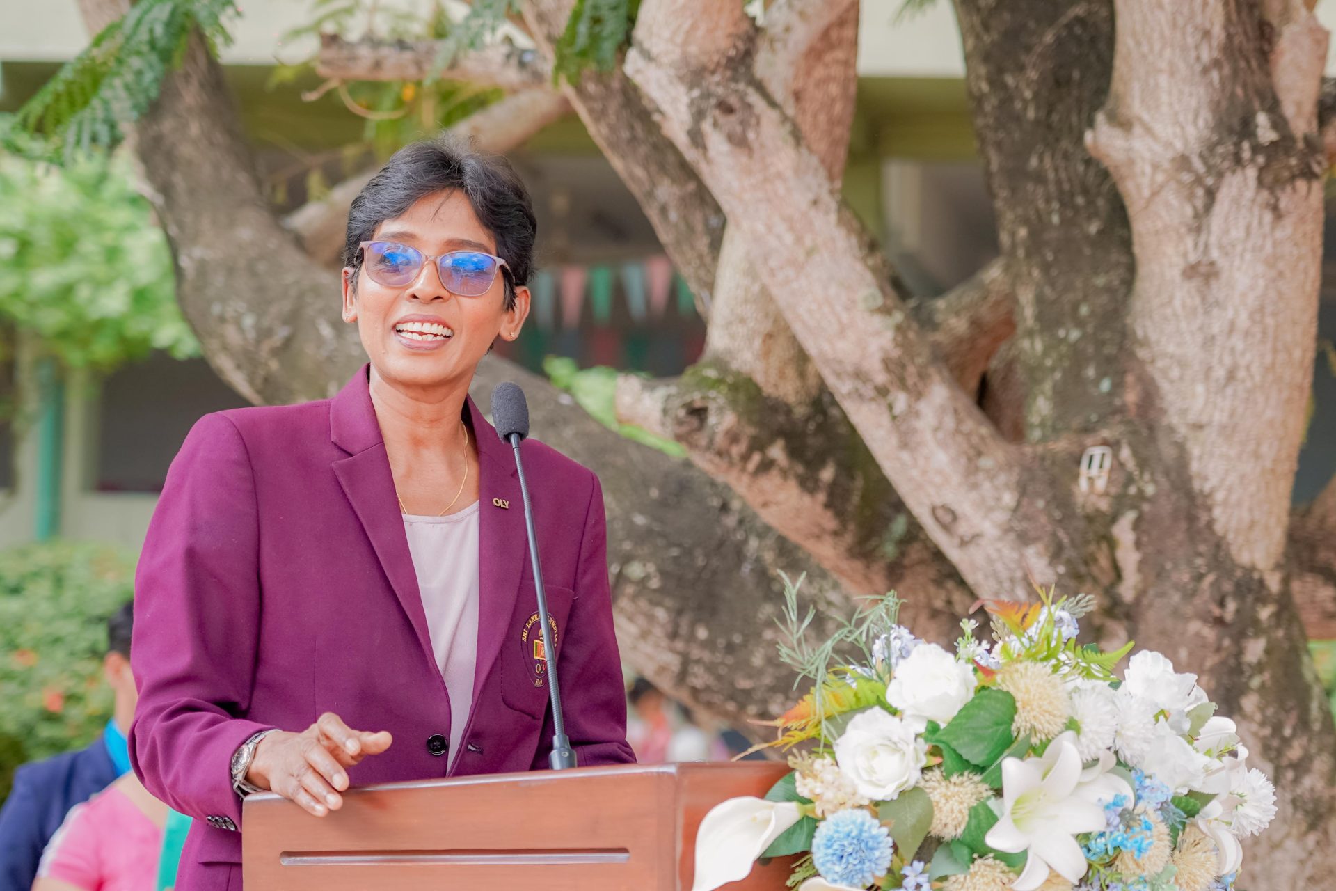 Person in maroon blazer speaks at outdoor podium with flowers, audience listening, tree in background.