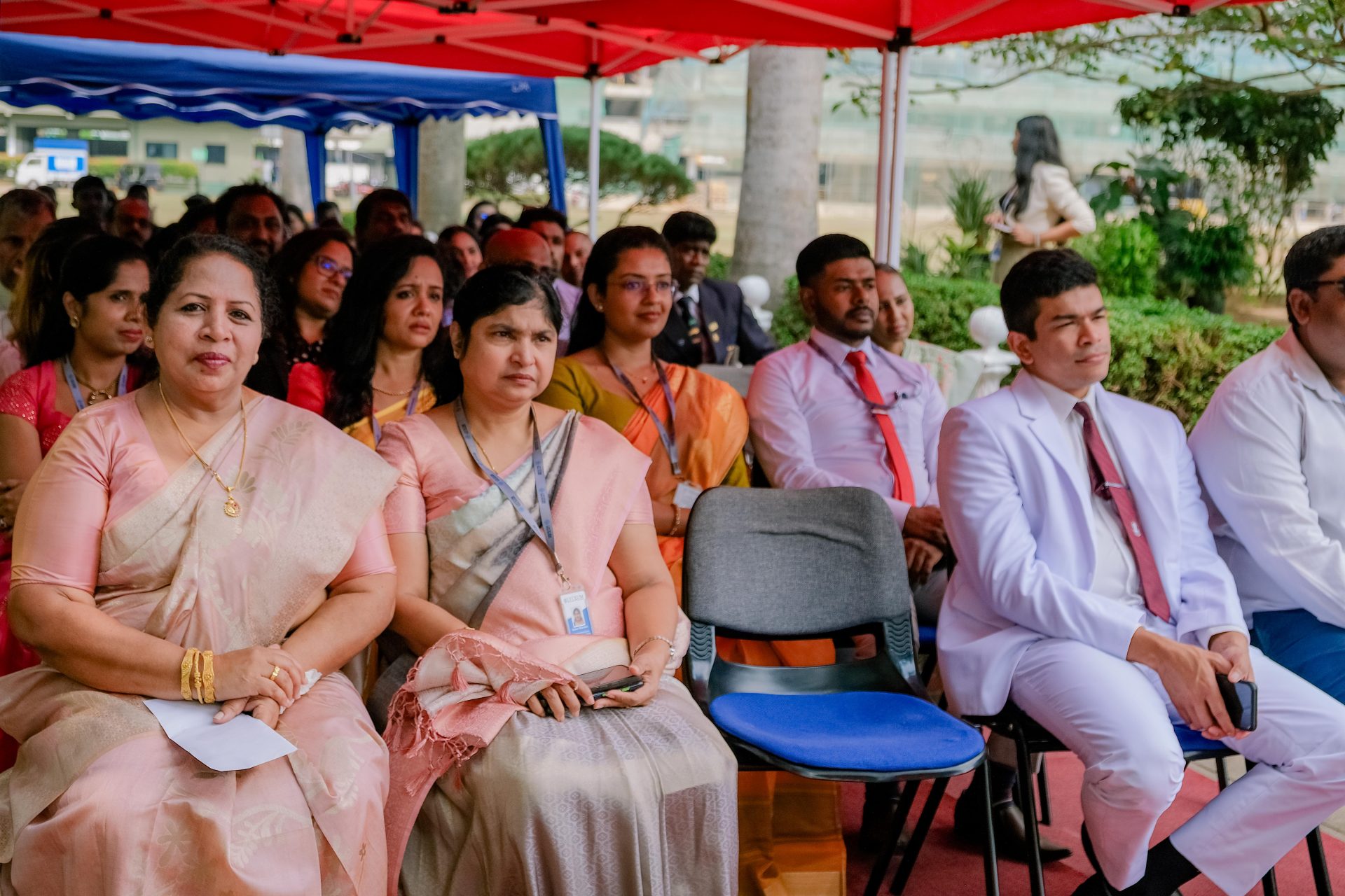 Diverse professionals in sarees and suits seated under red tents at formal outdoor ceremony.