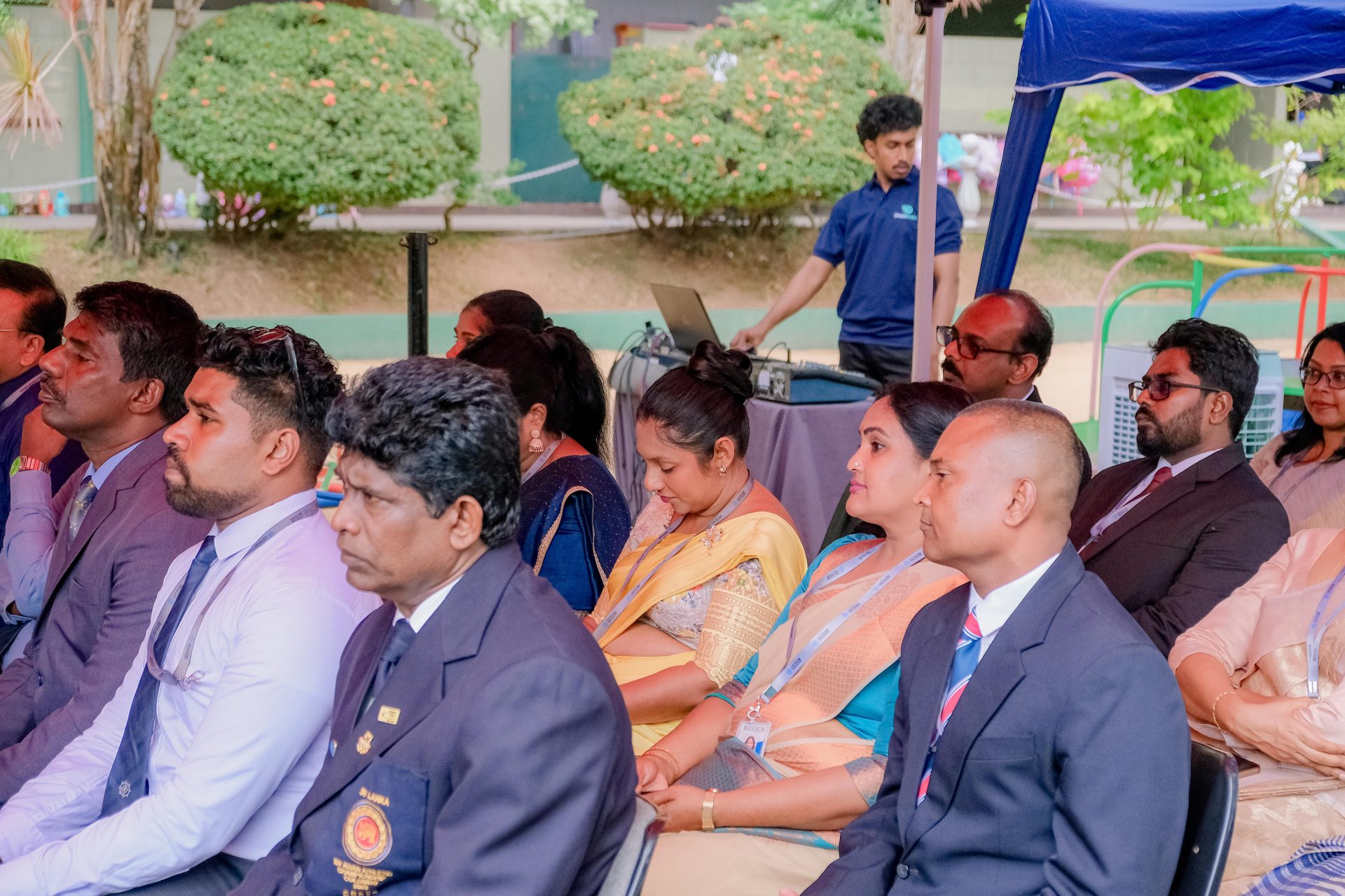 Professionals in suits and sarees seated outdoors at a formal ceremony with lush greenery.