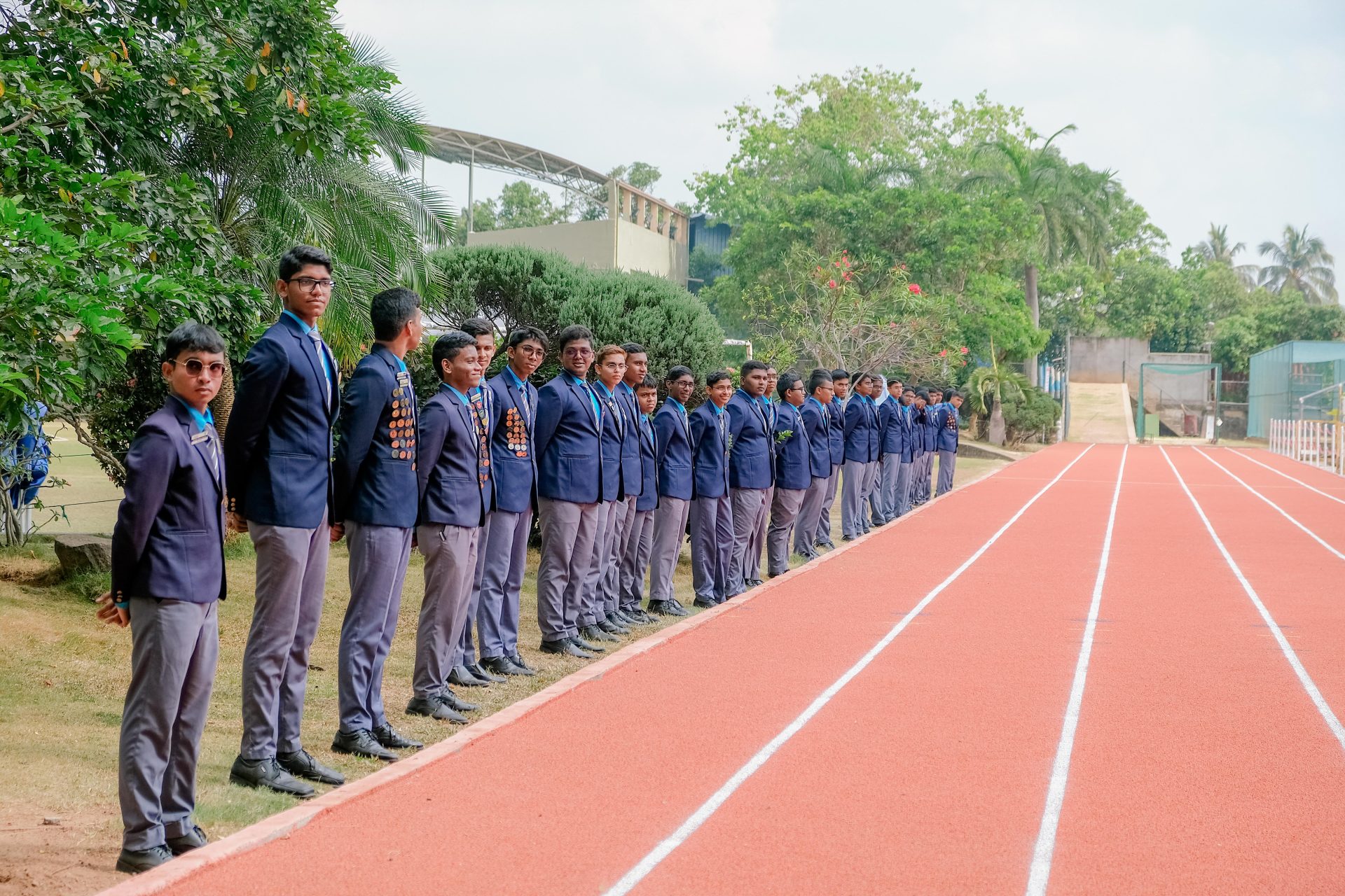 Schoolboys in uniform lined up by athletics track for outdoor ceremony, medals visible, tropical backdrop.
