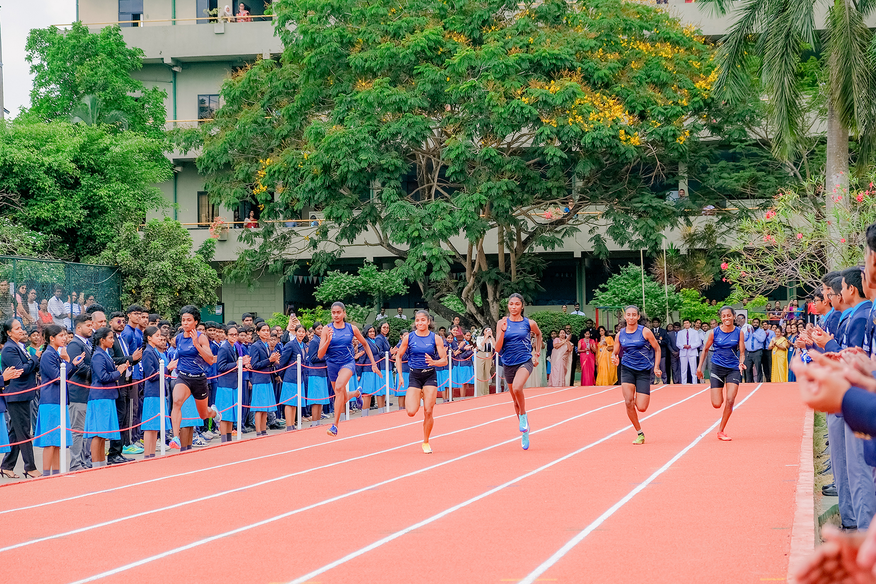 Schoolgirls sprinting on outdoor red track, cheered by classmates and teachers during school athletics event.